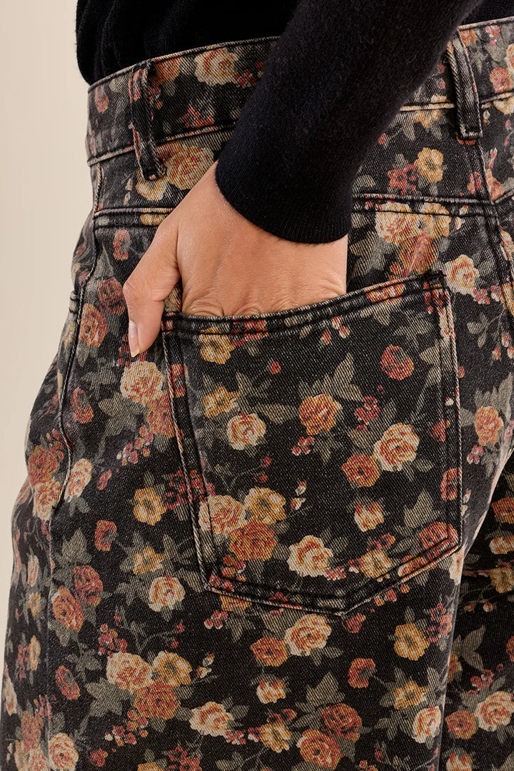 Close-up of a person wearing dark floral jeans with a hand in the back pocket, paired with a black long-sleeve top against a light beige background.