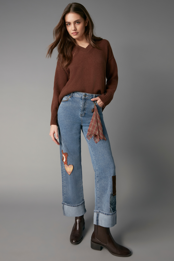 A model stands against a neutral gray studio background wearing a brown knit sweater, light-wash blue patchwork wide-leg jeans, and dark brown ankle boots.