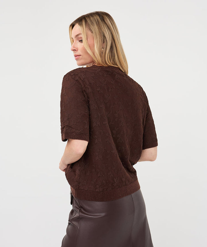 A woman shown from the back wearing a short-sleeve textured brown knit top and a dark brown mini skirt, photographed against a plain light gray studio background.