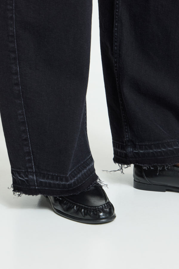 Close-up of a person wearing black wide-leg jeans with frayed hems and black leather loafers on a white studio background.