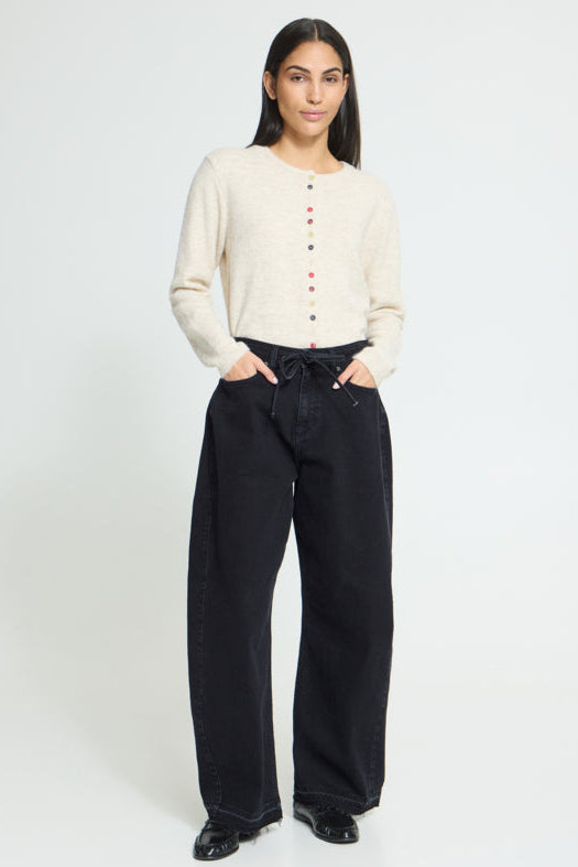 A woman modeling a cream ribbed knit cardigan with colorful buttons, paired with black wide-leg jeans and black shoes, against a plain white studio background.