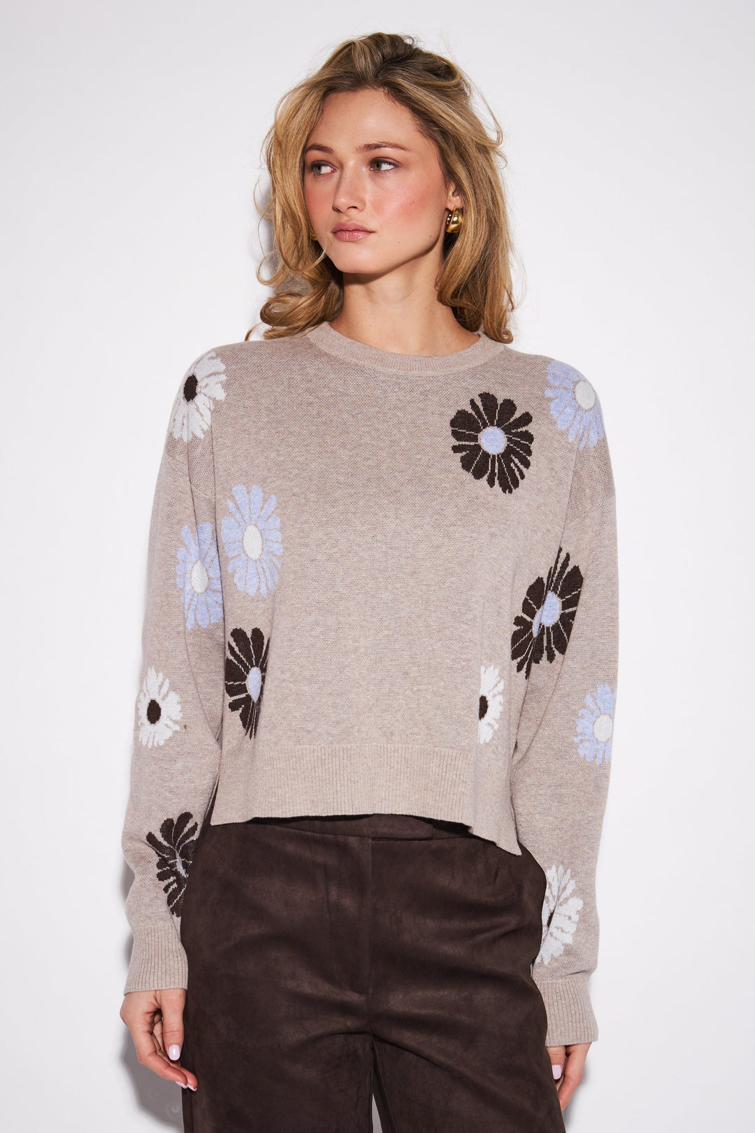 A woman modeling a cropped beige knit sweater with large black and white daisy patterns on the sleeves and shoulders, paired with dark brown wide-leg pants against a plain light gray studio background.