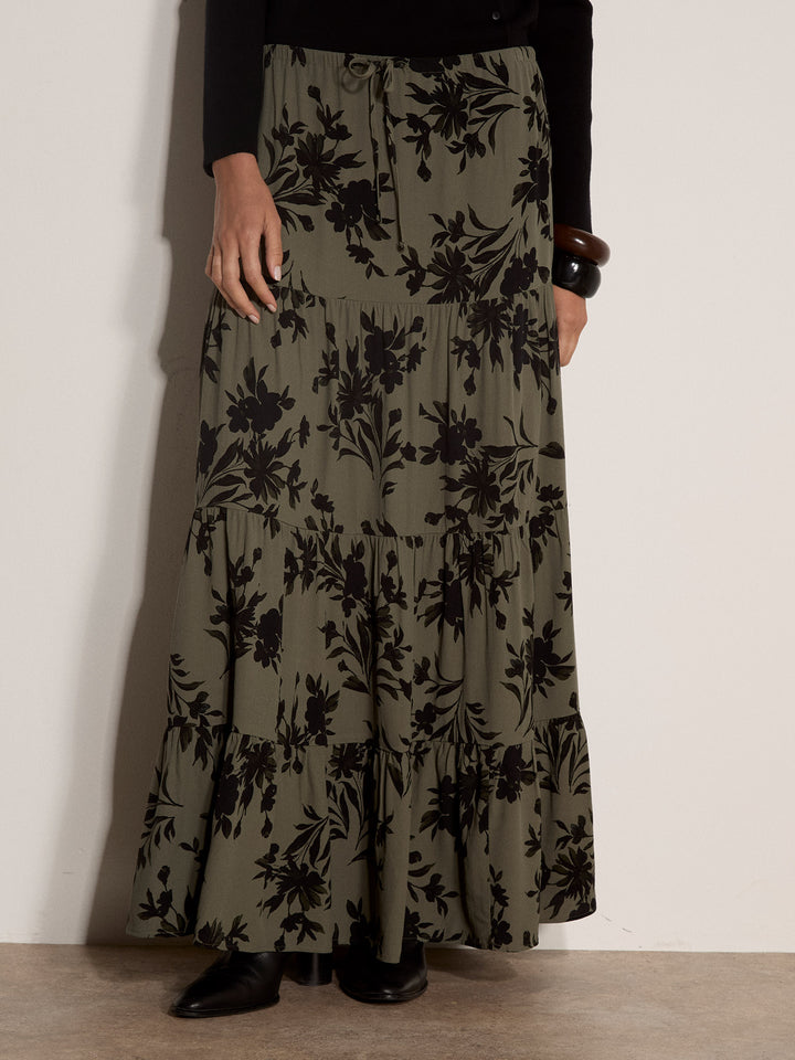 A model wearing a long, tiered maxi skirt in olive green with a black floral print, paired with a black top and black shoes, standing against a plain off-white wall.