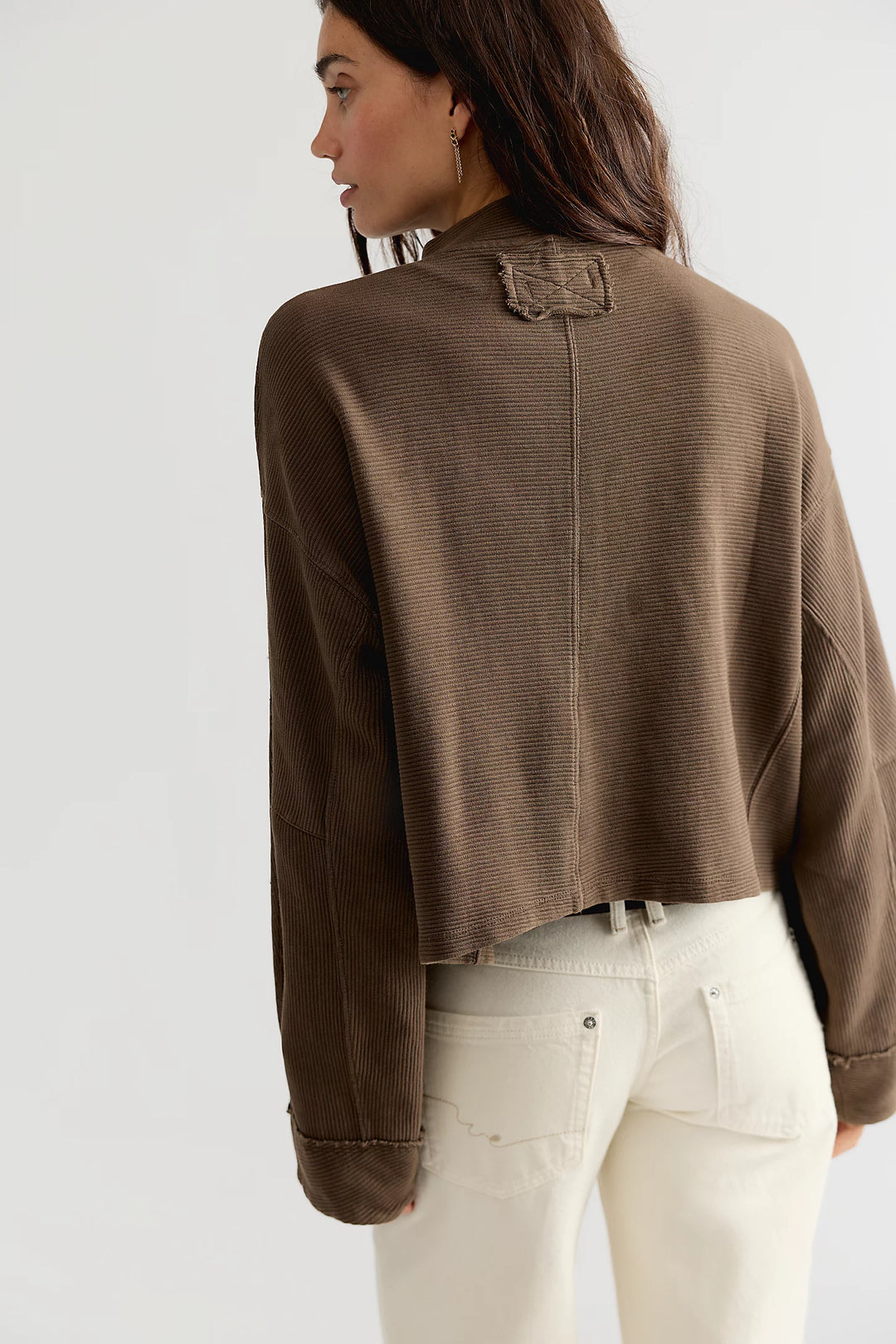 A woman shown from the back wearing a cropped brown textured knit sweater with a small decorative patch detail at the collar, paired with off-white jeans, against a plain light gray studio background.