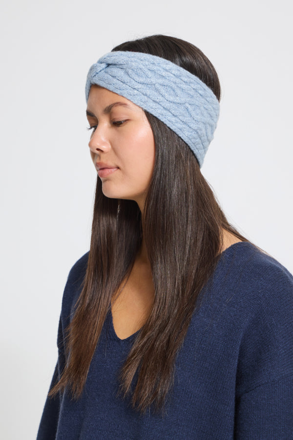 A woman wearing a light blue cable-knit twisted headband and a navy blue sweater, shown in profile against a plain light gray background.