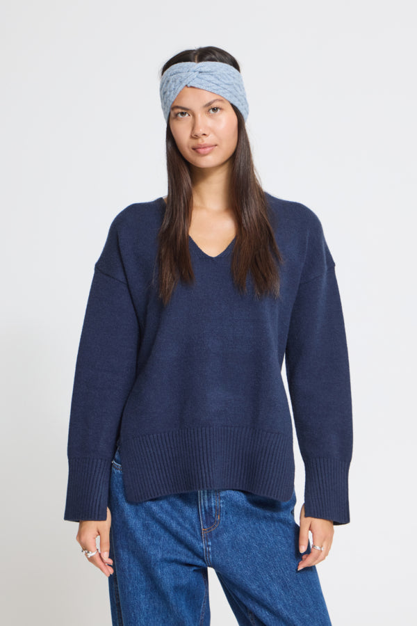 A woman wearing a navy blue knit V-neck sweater, blue jeans, and a light blue twisted headband, standing against a plain light gray studio background.