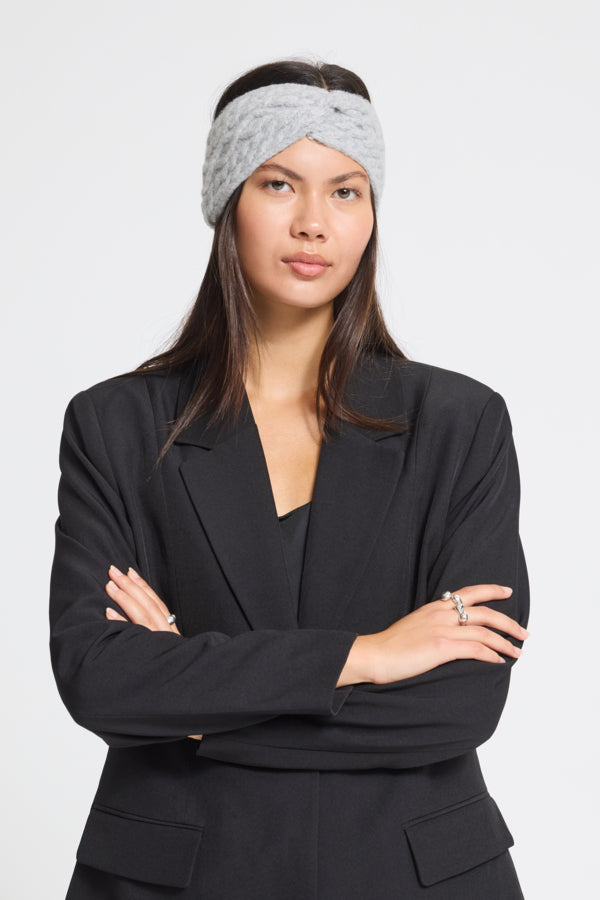 A woman wearing a light gray textured knit headband and a black blazer, posing with her arms crossed against a plain light gray background.