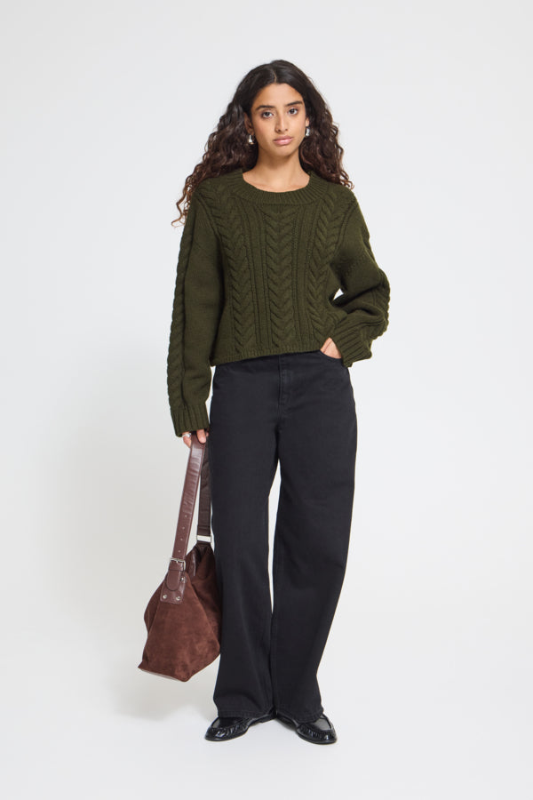 A woman stands against a light gray studio background wearing an olive green cable-knit cropped sweater, black wide-leg pants, and black shoes, while holding a brown shoulder bag.