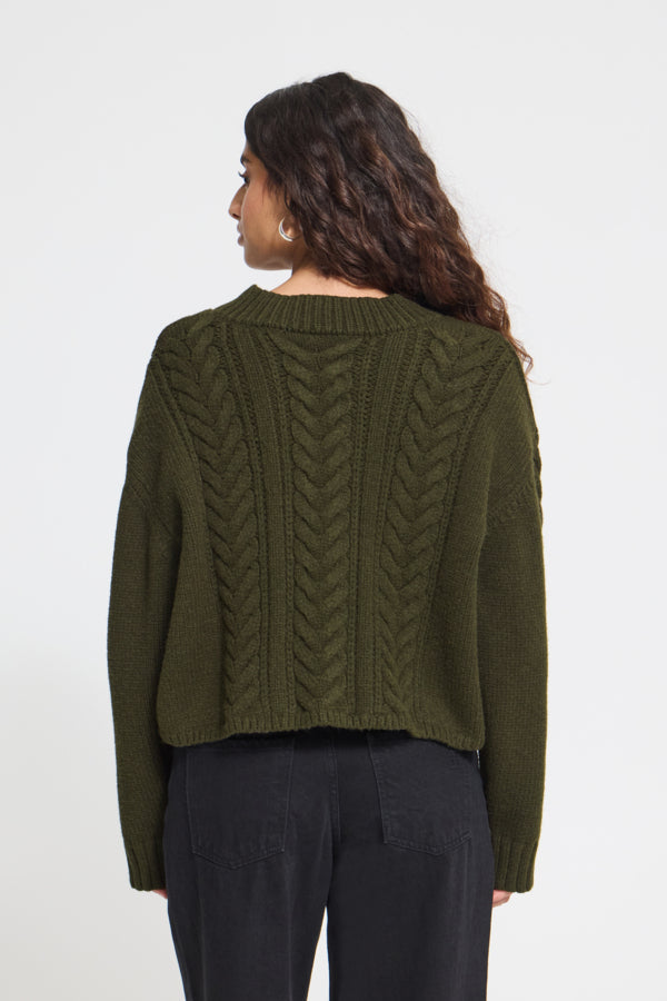 A woman viewed from the back wearing an olive green cable-knit cropped sweater and dark pants against a plain light gray studio background.