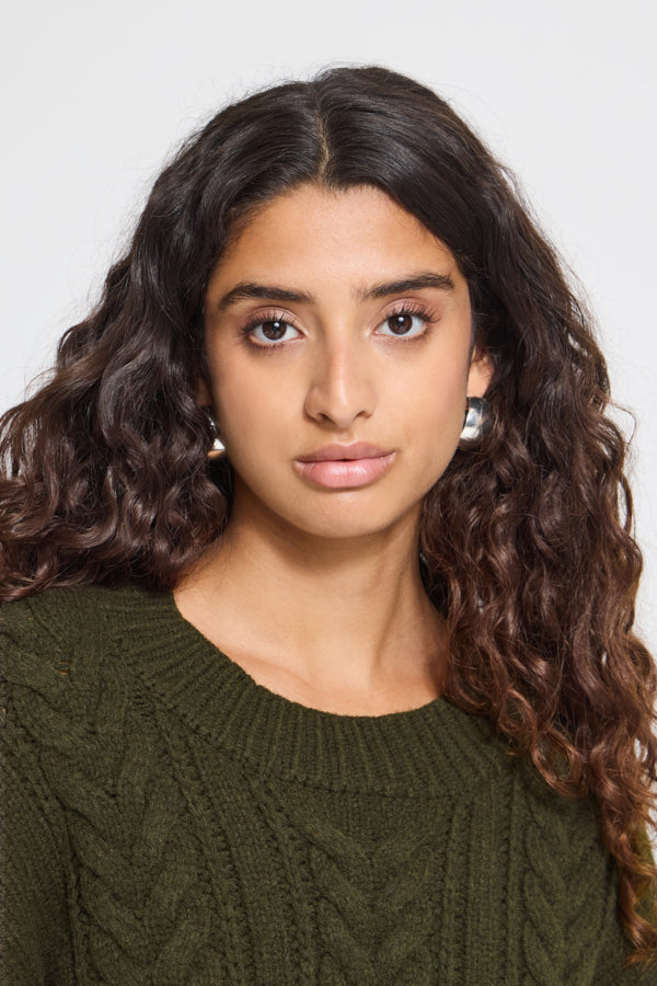 A woman with long, wavy dark brown hair wearing a textured olive green cable-knit sweater, shown from the chest up against a light gray studio background.