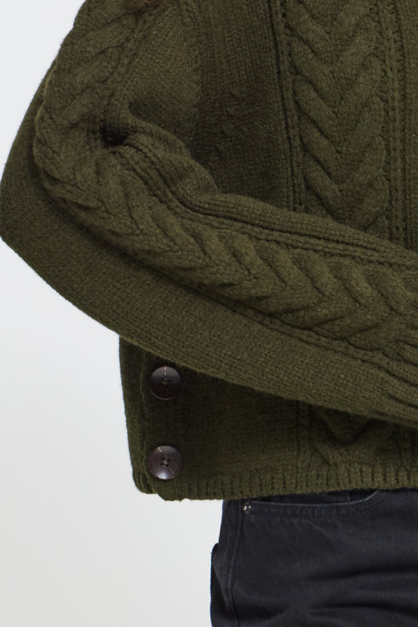 Close-up of an olive green cable-knit sweater with dark buttons, worn with dark navy pants against a light gray background.