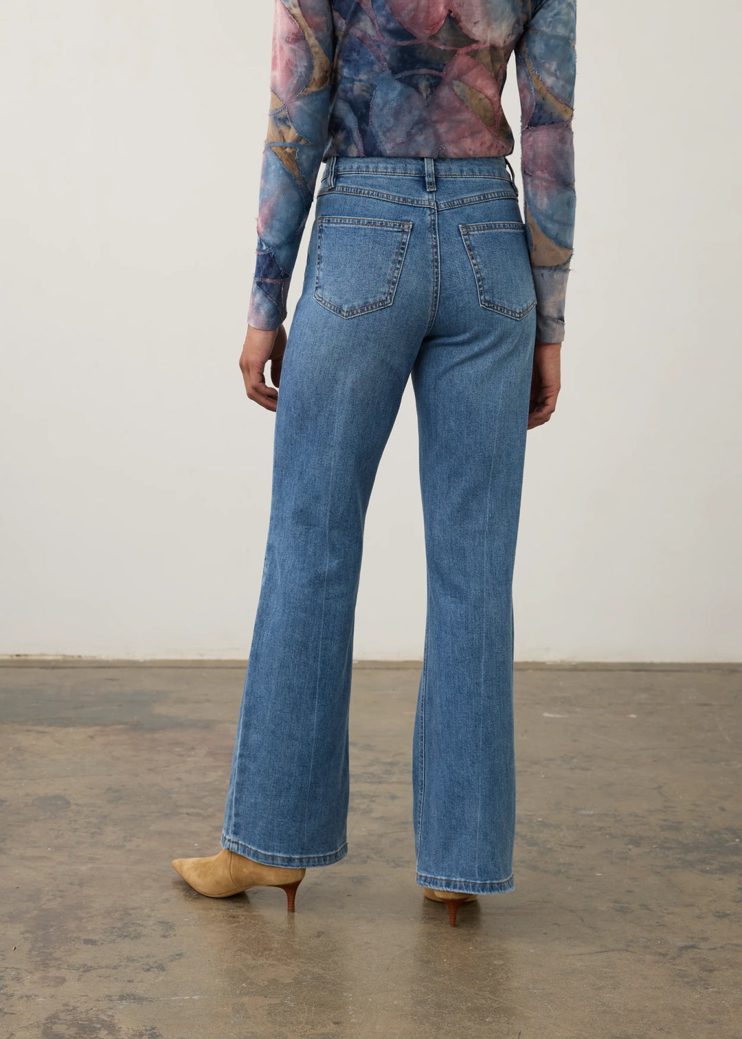 A person shown from the back wearing blue wide-leg jeans, a long-sleeve floral top, and tan pointed-toe heels against a plain indoor wall and concrete floor.
