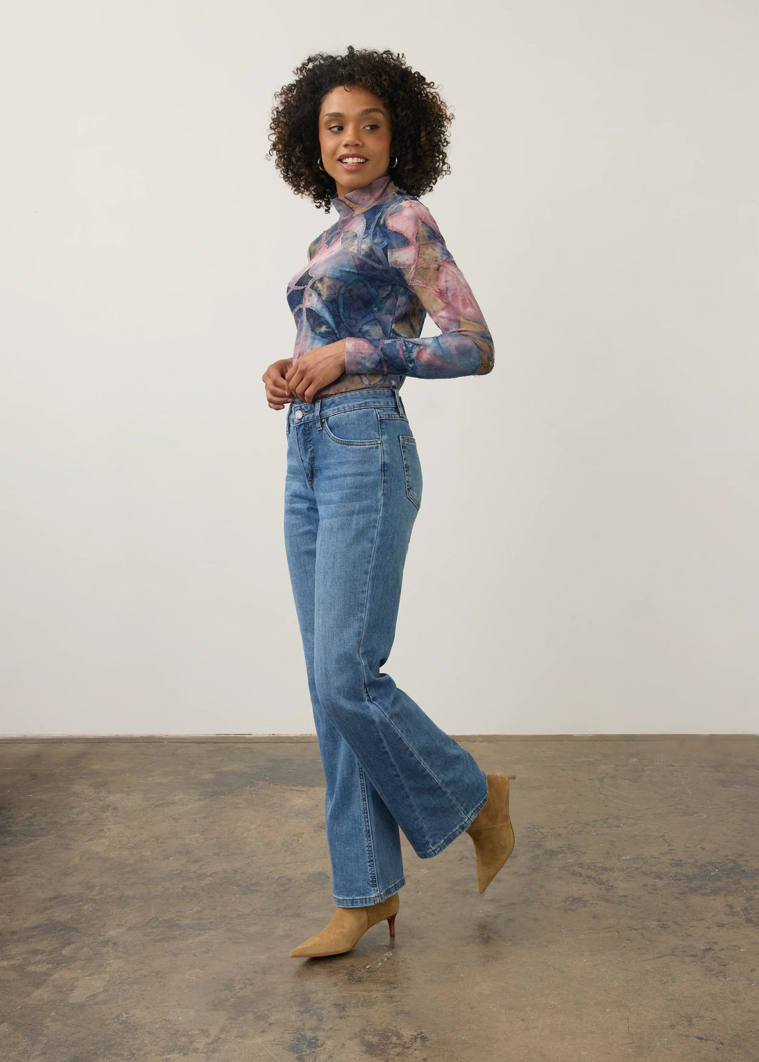 A woman modeling a sheer long-sleeve top with a blue, pink, and purple floral pattern, paired with high-waisted blue flare jeans and tan pointed-toe heels, standing against a plain off-white wall and concrete floor.