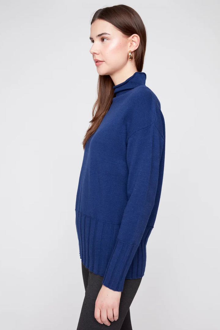A woman shown in side profile wearing a navy blue ribbed turtleneck sweater and black pants against a light gray studio background.