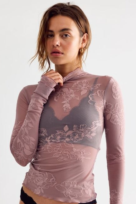 A woman modeling a sheer, long-sleeve high-neck top in dusty mauve with tonal floral lace patterns and a smocked chest panel, paired with black lace panties against a plain light gray background.