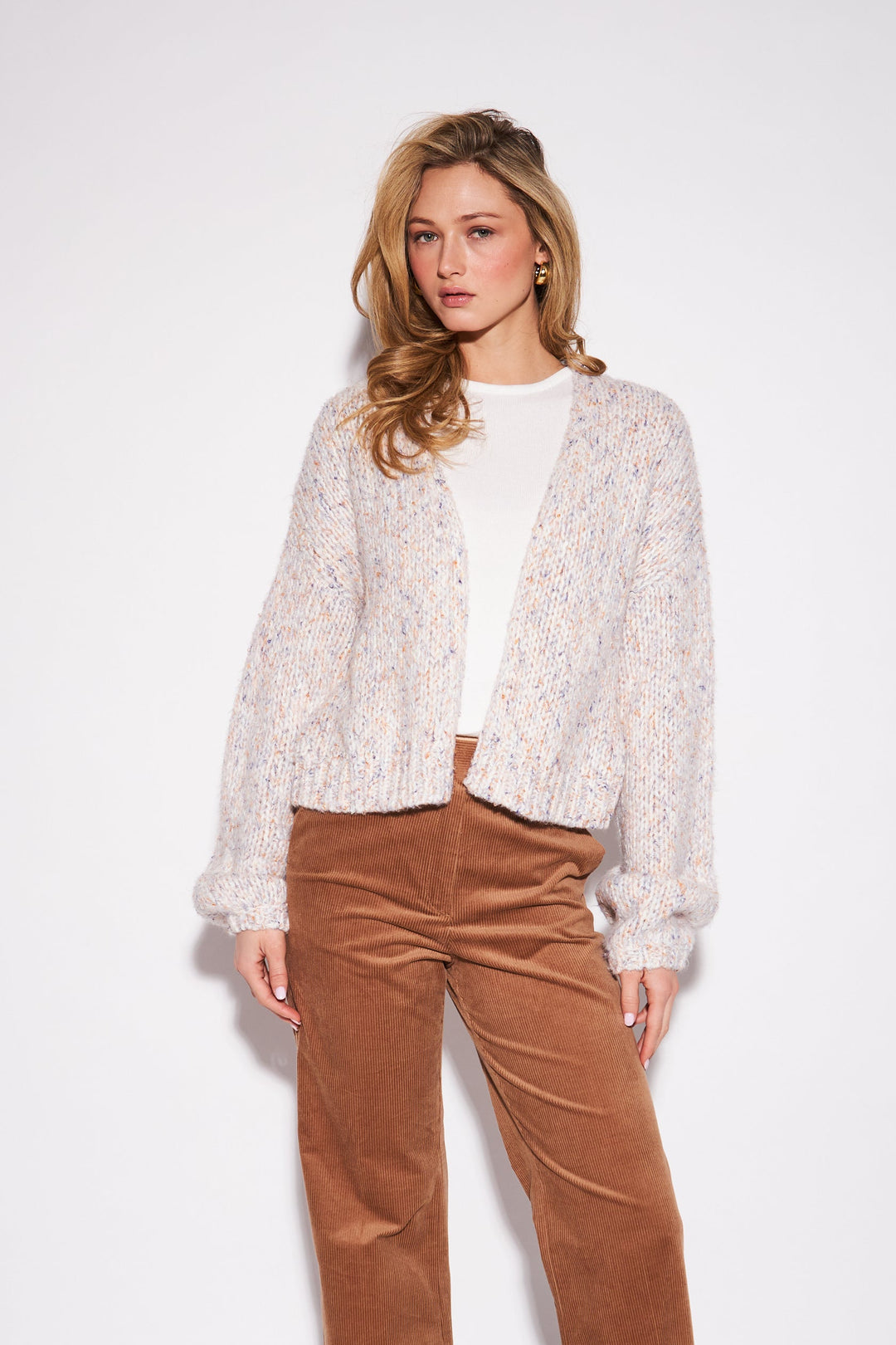 A woman stands against a plain light gray studio background wearing a white textured knit cardigan over a white top, brown wide-leg corduroy pants, and dark brown pointed heels.