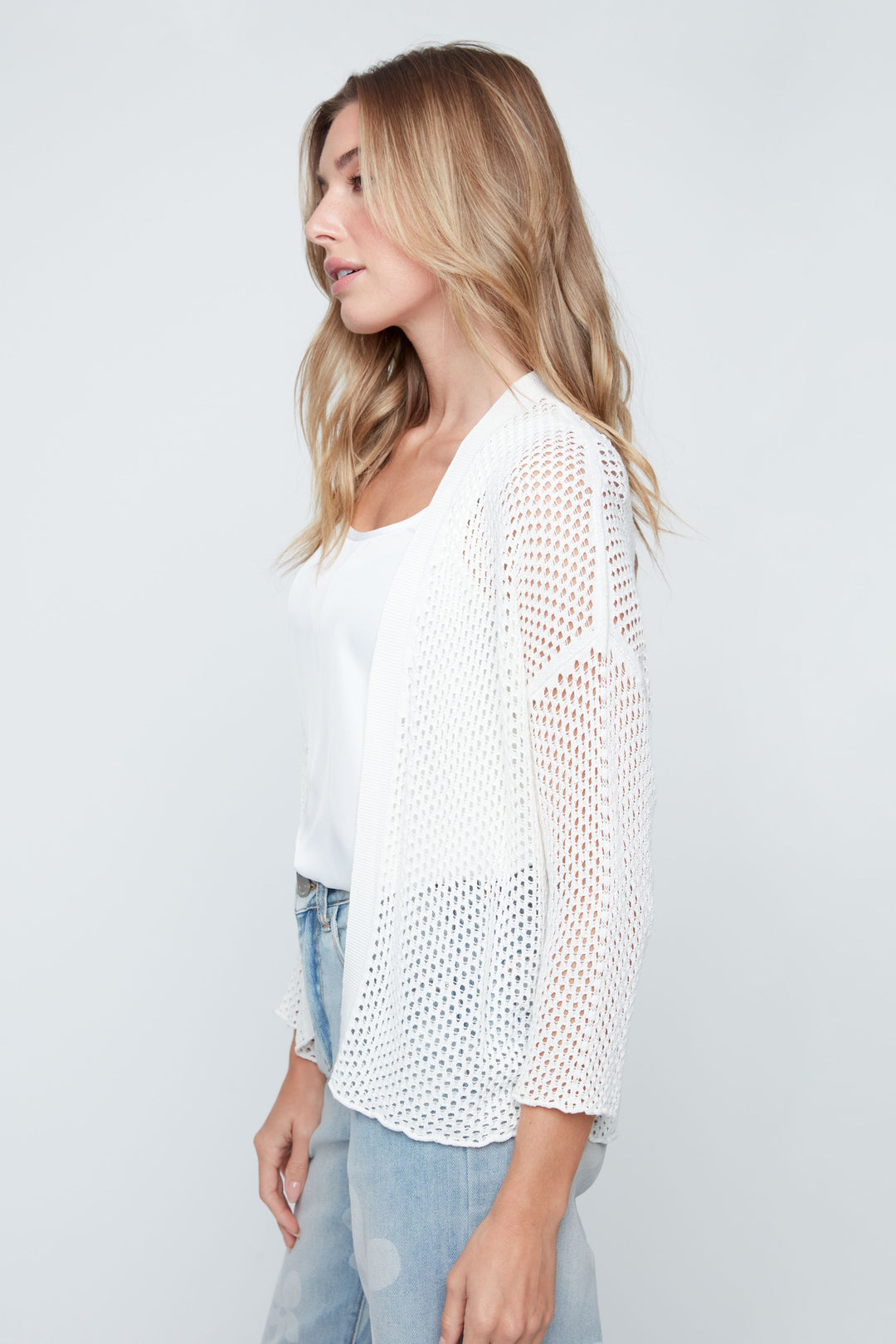 Woman wearing a white knit cardigan over a white shirt with light blue jeans on a plain background