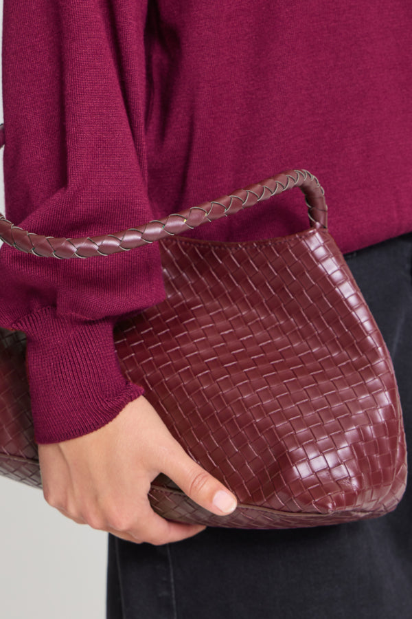A close-up of a person wearing a burgundy knit sweater and holding a woven leather shoulder bag in a deep burgundy-brown shade. The bag has a structured, curved silhouette and a matching woven handle.
