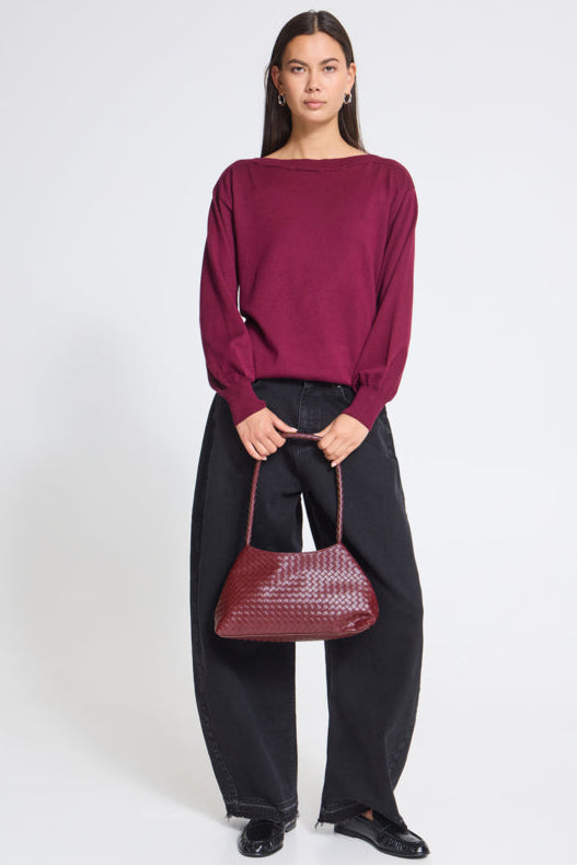 A model stands against a light gray studio background wearing a deep burgundy long-sleeve knit top, black wide-leg pants, and black loafers, while holding a matching burgundy woven handbag.