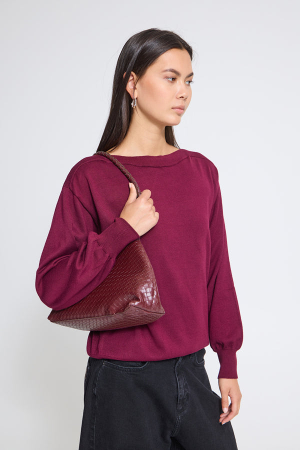 A woman wearing a maroon long-sleeve knit top and black pants, carrying a maroon textured crocodile-embossed shoulder bag against a light gray studio background.