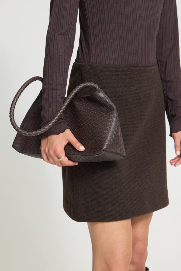 A person wearing a dark brown ribbed long-sleeve top and a dark brown skirt holds a dark brown woven leather handbag with a thick braided handle against a light gray studio background.