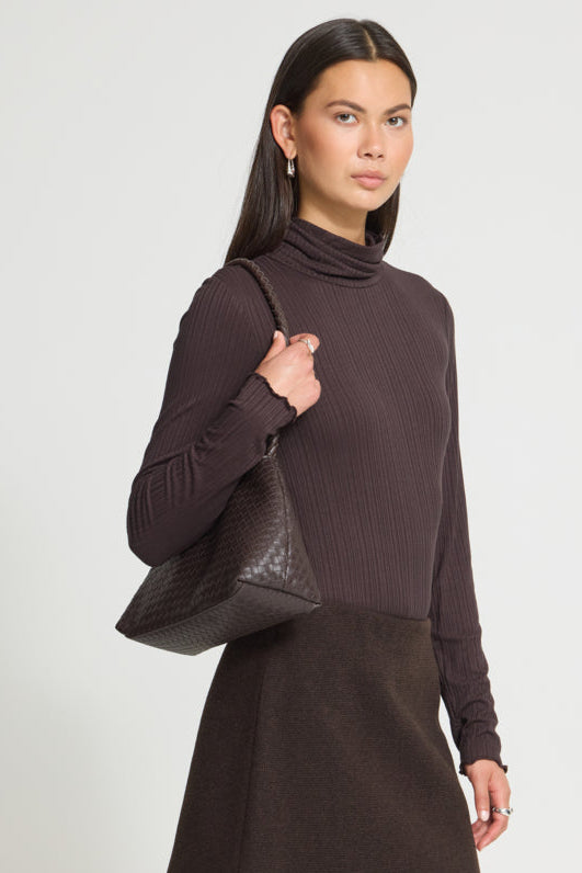A woman modeling a dark brown ribbed turtleneck sweater and matching dark brown skirt, carrying a dark brown woven leather shoulder bag against a light gray studio background.