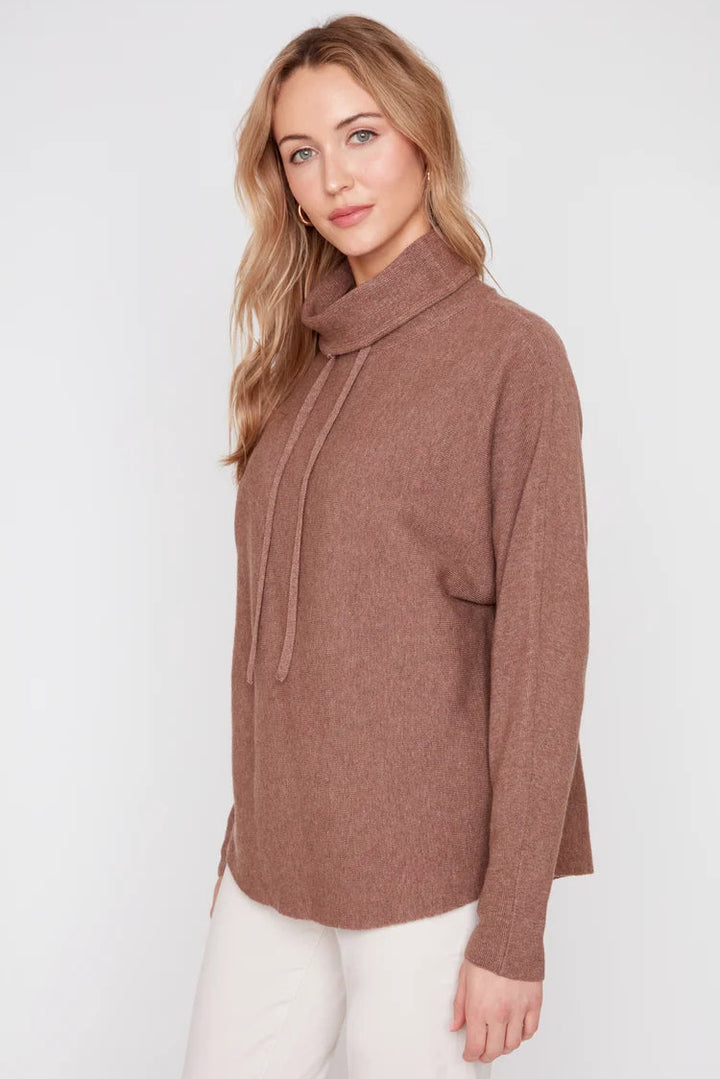 A woman modeling a brown knit cowl-neck sweater with a drawstring and front pocket, paired with white pants, against a plain light gray studio background.
