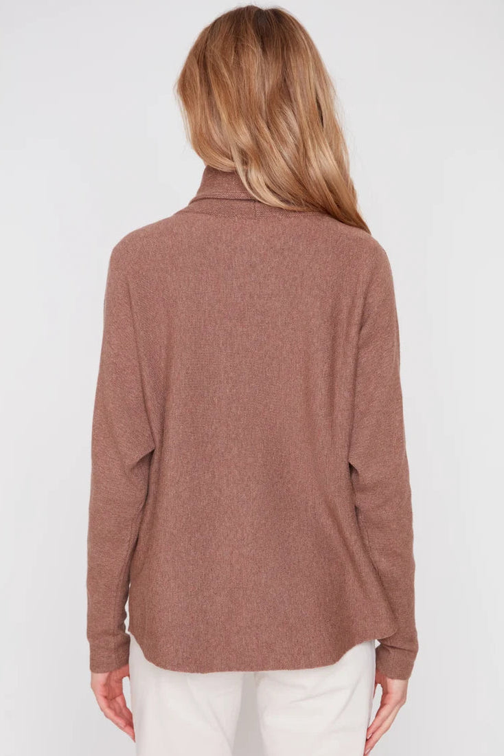 A woman viewed from the back wearing a long-sleeve, high-neck knit top in a dusty rose-brown color, paired with white pants, against a plain light gray studio background.