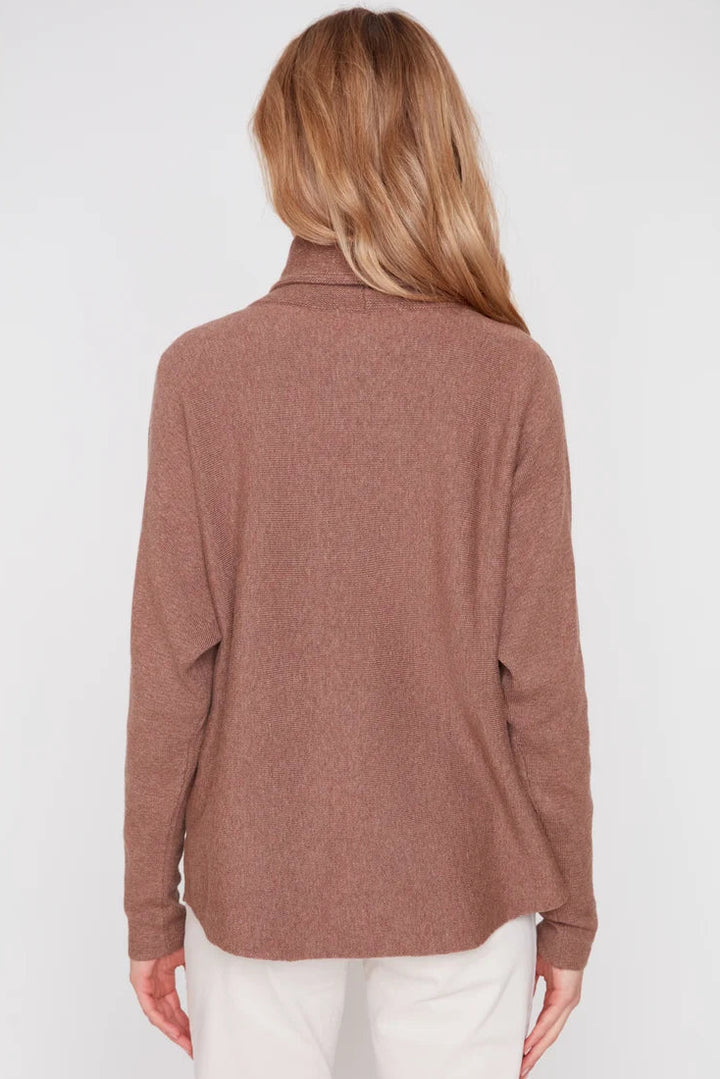 A woman viewed from the back wearing a long-sleeve, high-neck knit top in a dusty rose-brown color, paired with white pants, against a plain light gray studio background.