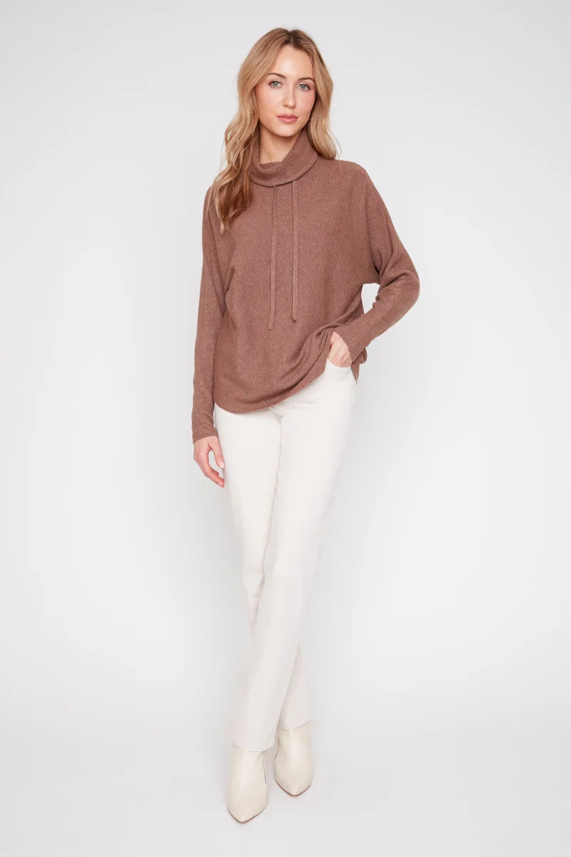 A woman modeling a brown long-sleeve knit cowl-neck sweater with drawstrings, paired with off-white pants and cream ankle boots, against a plain light gray studio background.