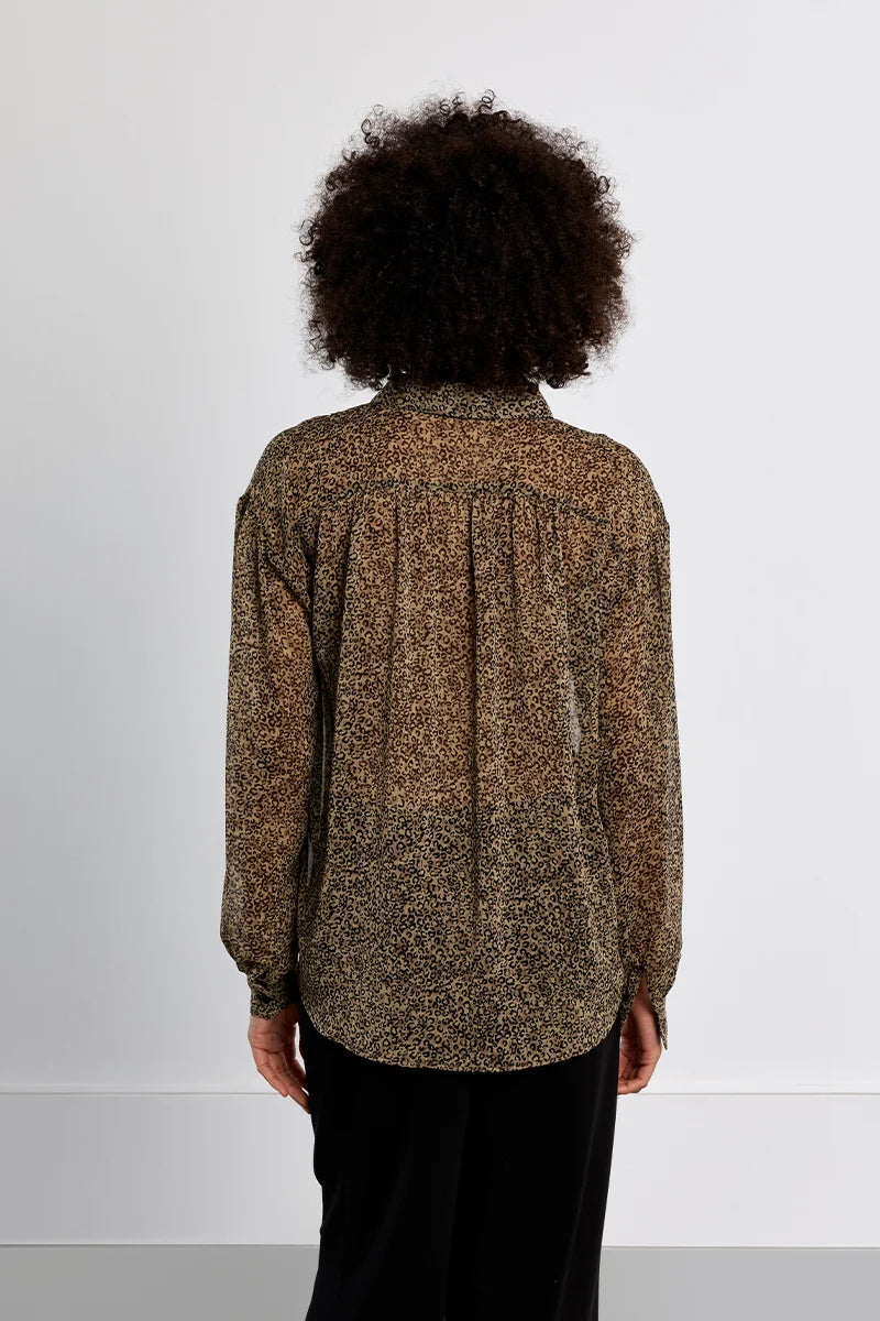 A person viewed from the back wearing a long-sleeve blouse with a mottled brown and beige pattern and black pants, standing against a plain light gray wall.
