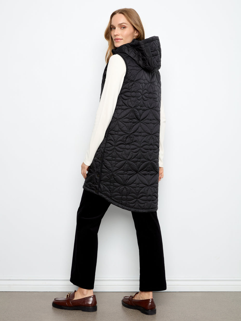 A woman modeling a black quilted hooded sleeveless puffer vest over a white long-sleeve top, black trousers, and brown loafers, standing against a plain white wall.