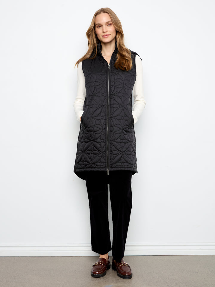 A woman modeling a black quilted sleeveless zip-front vest over a white long-sleeve top, black pants, and brown loafers, standing against a plain white wall.