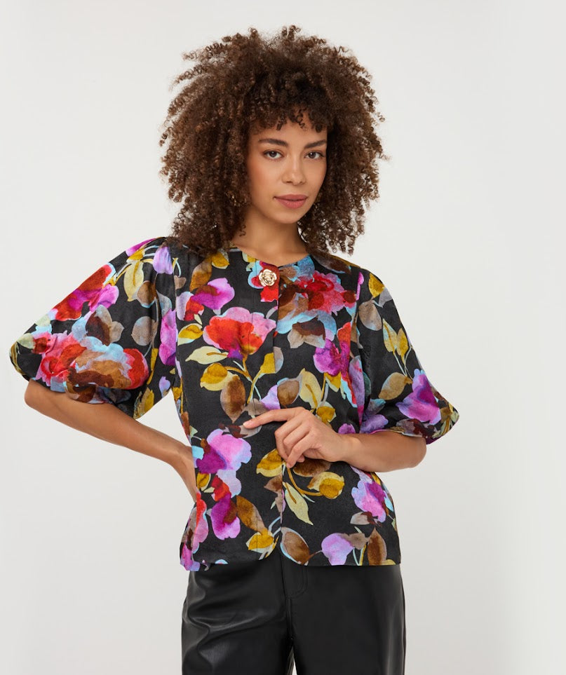 A woman with curly brown hair wearing a black short-sleeve blouse with a vibrant multicolor floral print and puff sleeves, paired with black pants, against a plain light gray background.