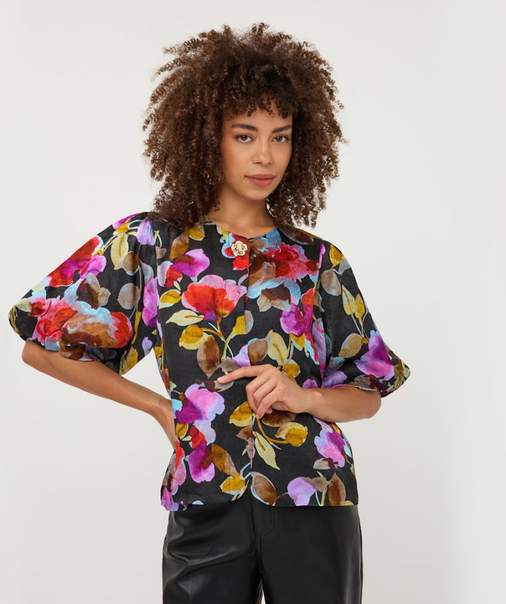 A woman with curly brown hair wearing a black short-sleeve blouse with a vibrant multicolor floral print and puff sleeves, paired with black pants, against a plain light gray background.