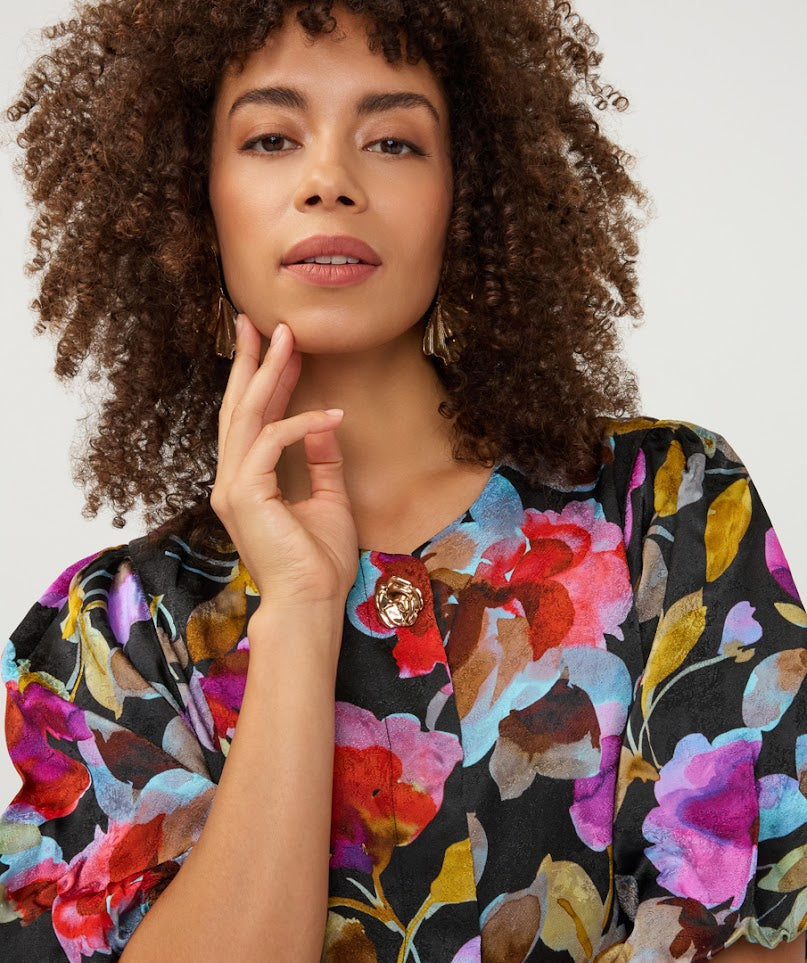 A close-up of a woman with curly brown hair wearing a black blouse with a vibrant multicolor floral print and a decorative gold button at the neckline, set against a plain light background.