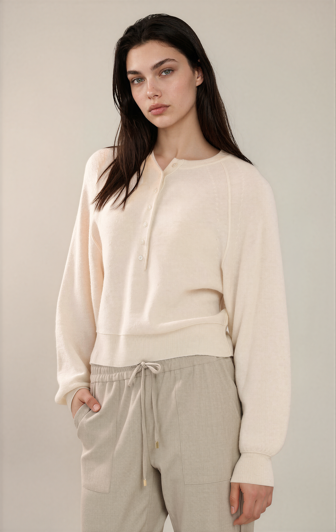 A woman modeling a cream-colored long-sleeve knit henley top paired with light beige drawstring trousers, set against a plain beige studio background.