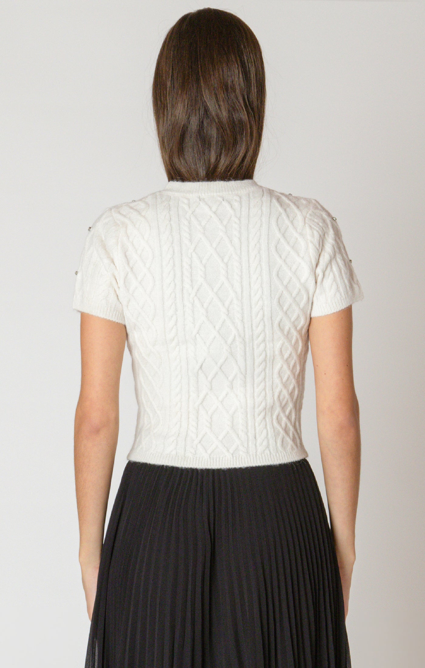 Person wearing a white cable knit top and black pleated skirt on a plain background