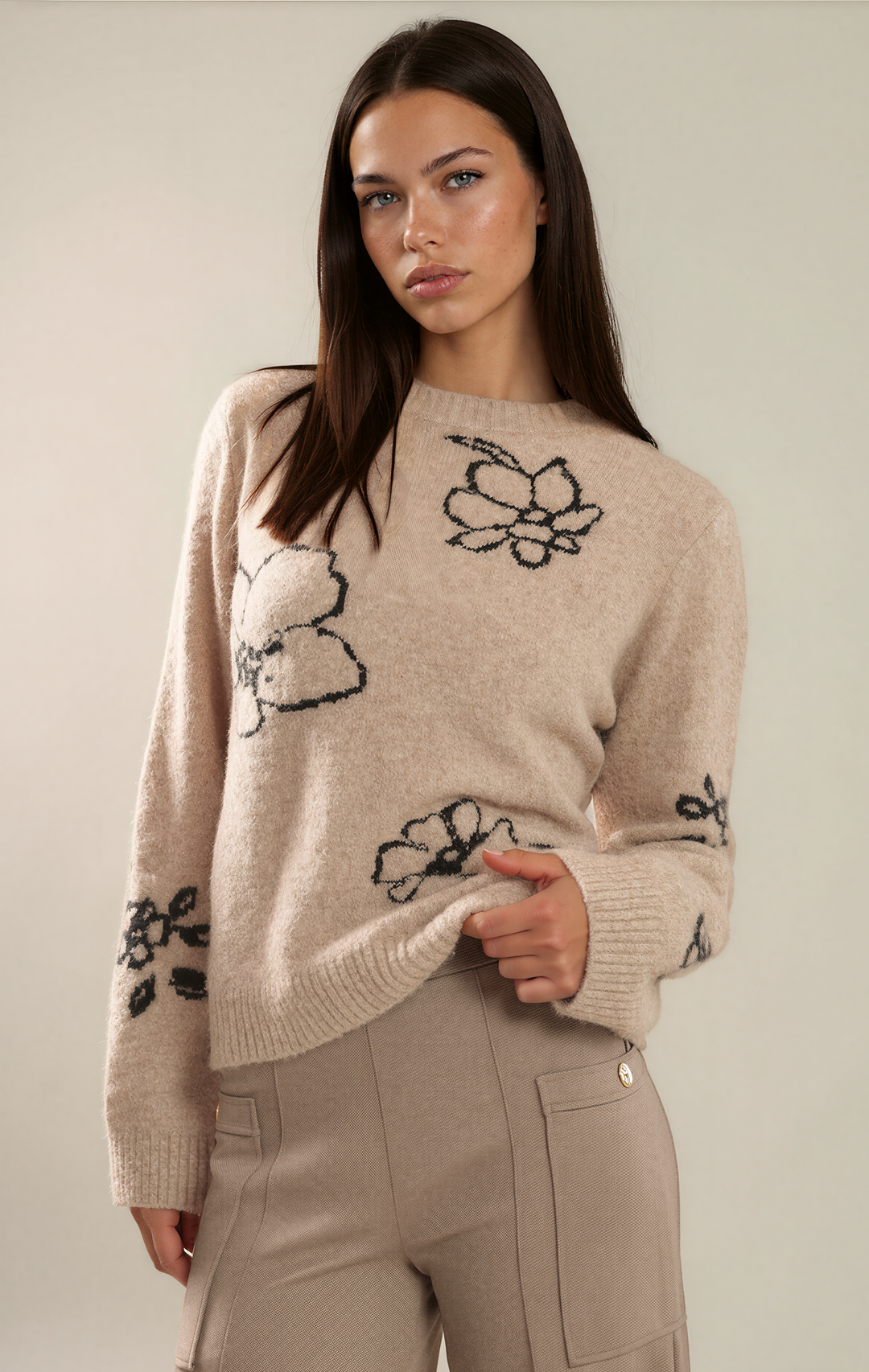 A woman modeling a beige knit sweater with black floral motifs and matching beige trousers against a plain light beige studio background.