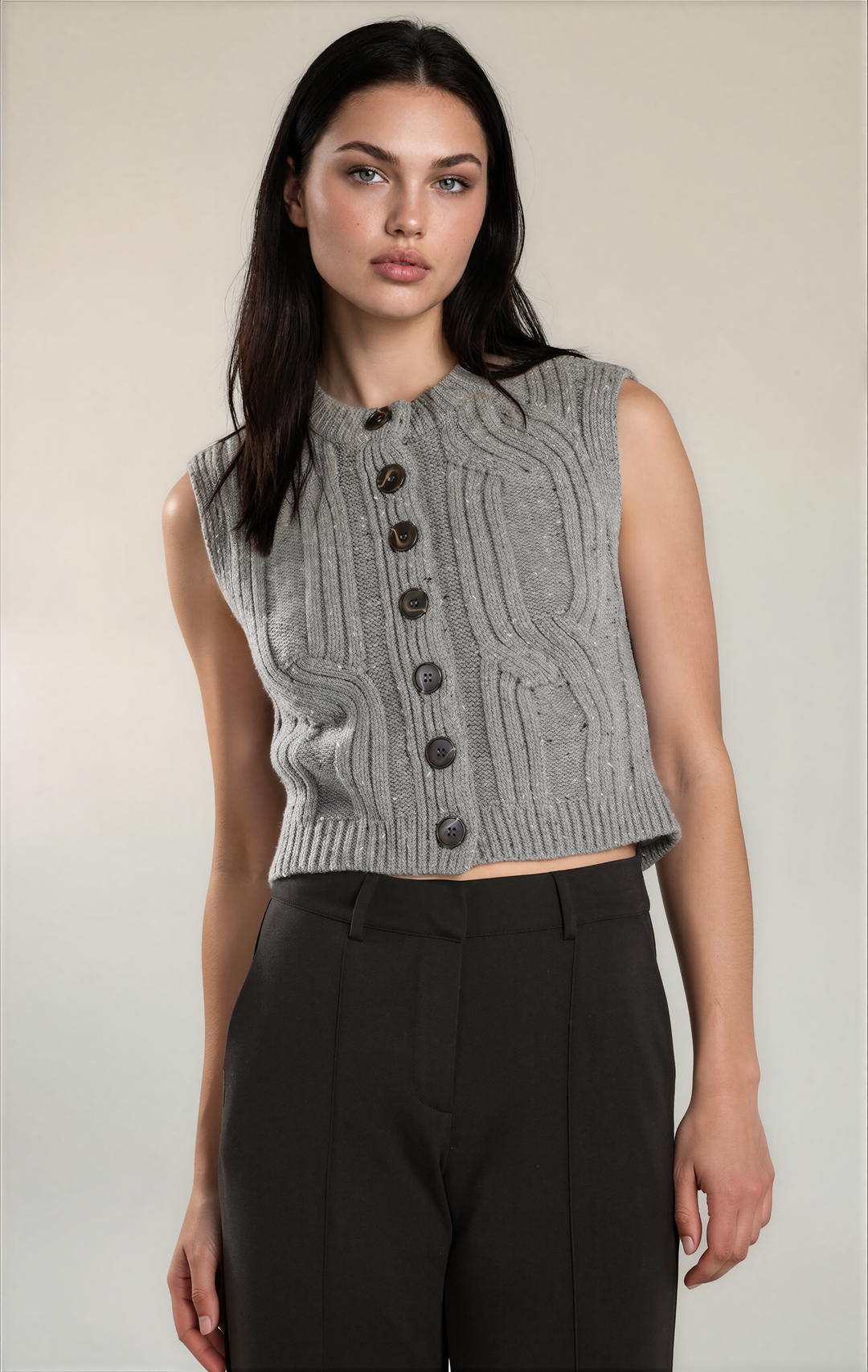 A woman modeling a sleeveless gray cable-knit cropped vest with dark buttons, paired with black trousers, against a plain light beige studio background.