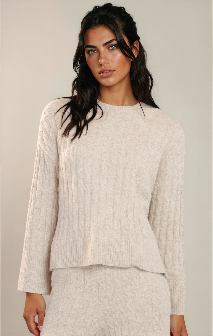 A woman modeling a cream-colored cable-knit crewneck sweater with matching textured knit bottoms against a plain light gray studio background.
