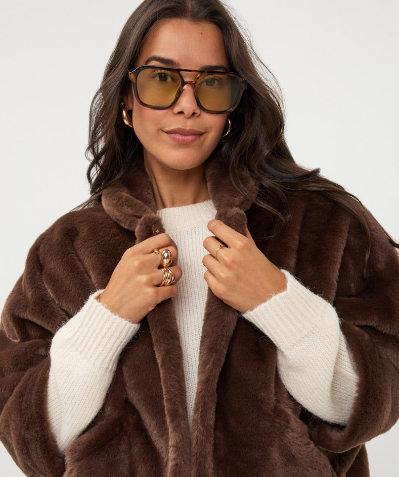 A woman wearing a plush brown faux fur coat over a cream ribbed sweater, accessorized with gold hoop earrings and gold-framed sunglasses, against a plain light gray background.