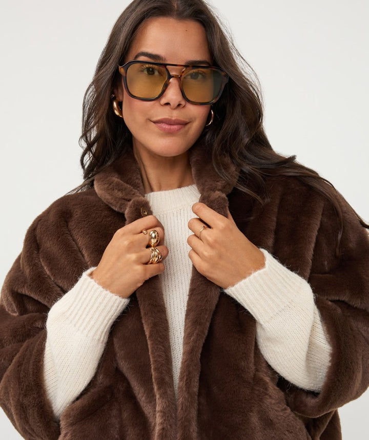 A woman wearing a plush brown faux fur coat over a cream ribbed sweater, accessorized with gold hoop earrings and gold-framed sunglasses, against a plain light gray background.