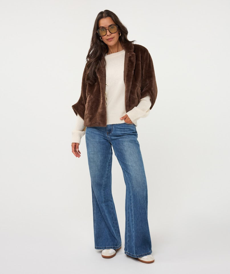 A woman modeling a brown faux fur jacket over a cream knit top and blue wide-leg jeans, standing against a plain white studio background.