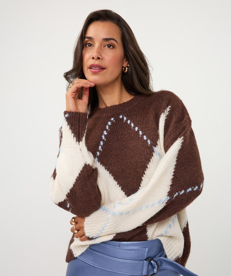 A woman wearing a brown and cream argyle knit sweater with light blue dotted accents, paired with blue leather pants, against a plain light gray background.