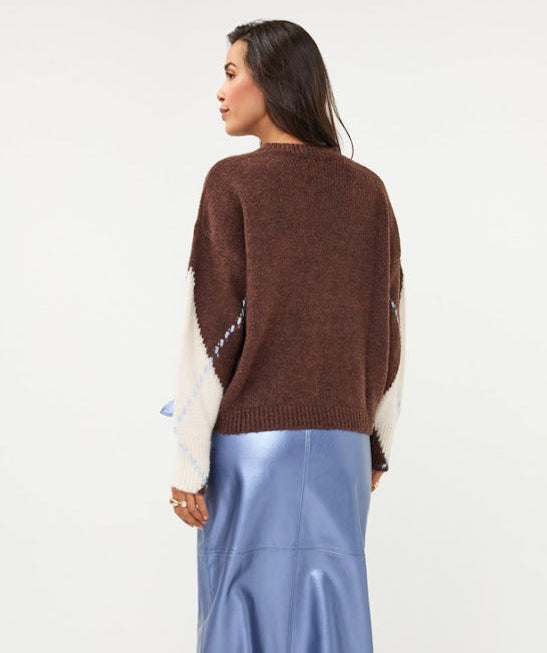 A woman is shown from the back wearing a brown knit sweater with cream-colored side panels, a light blue satin midi skirt, and dark brown heeled boots, set against a plain white studio background.