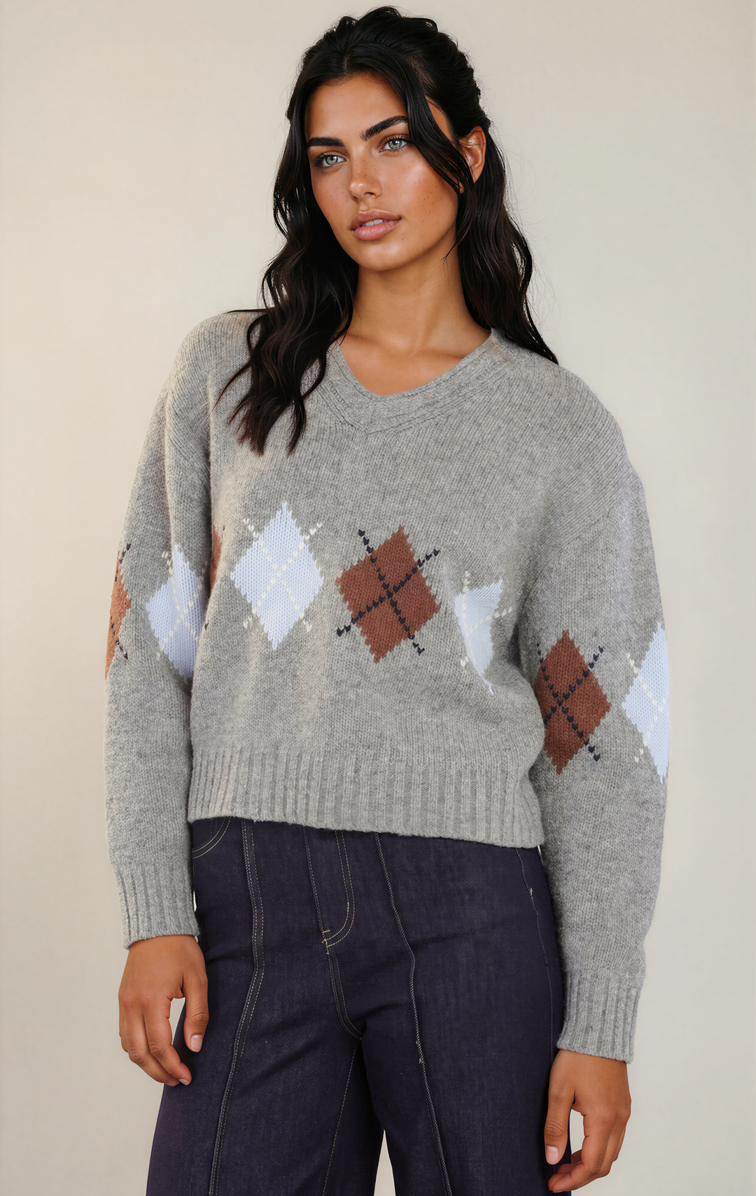 A woman wearing a light gray knit sweater with an argyle pattern in white, brown, and dark brown, paired with dark blue jeans, against a plain beige background.