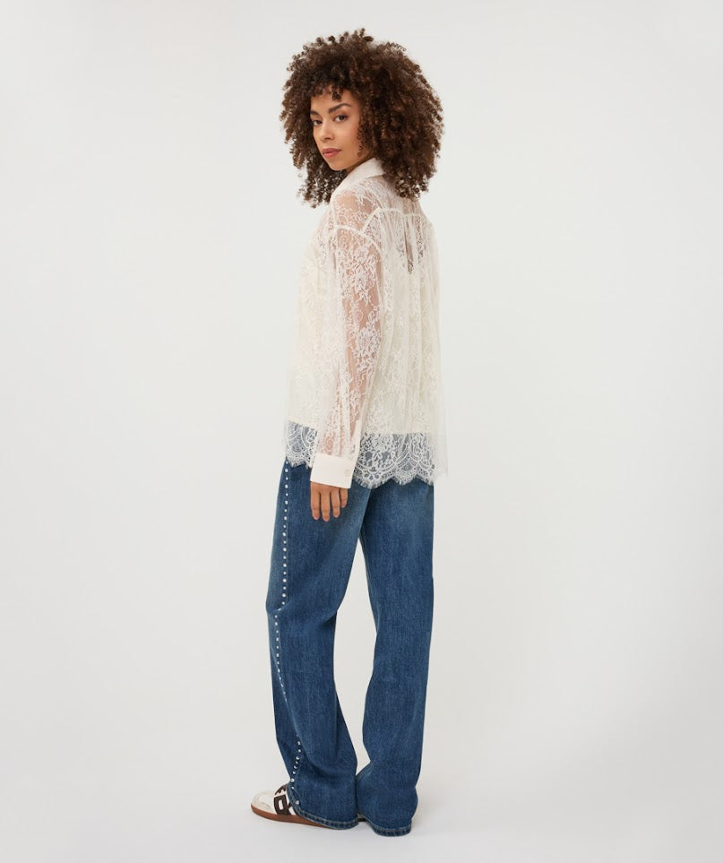 A model with curly brown hair is shown from a side-back angle wearing a sheer white lace long-sleeve top with scalloped edges and blue jeans with silver stud detailing down the side, set against a plain white studio background.