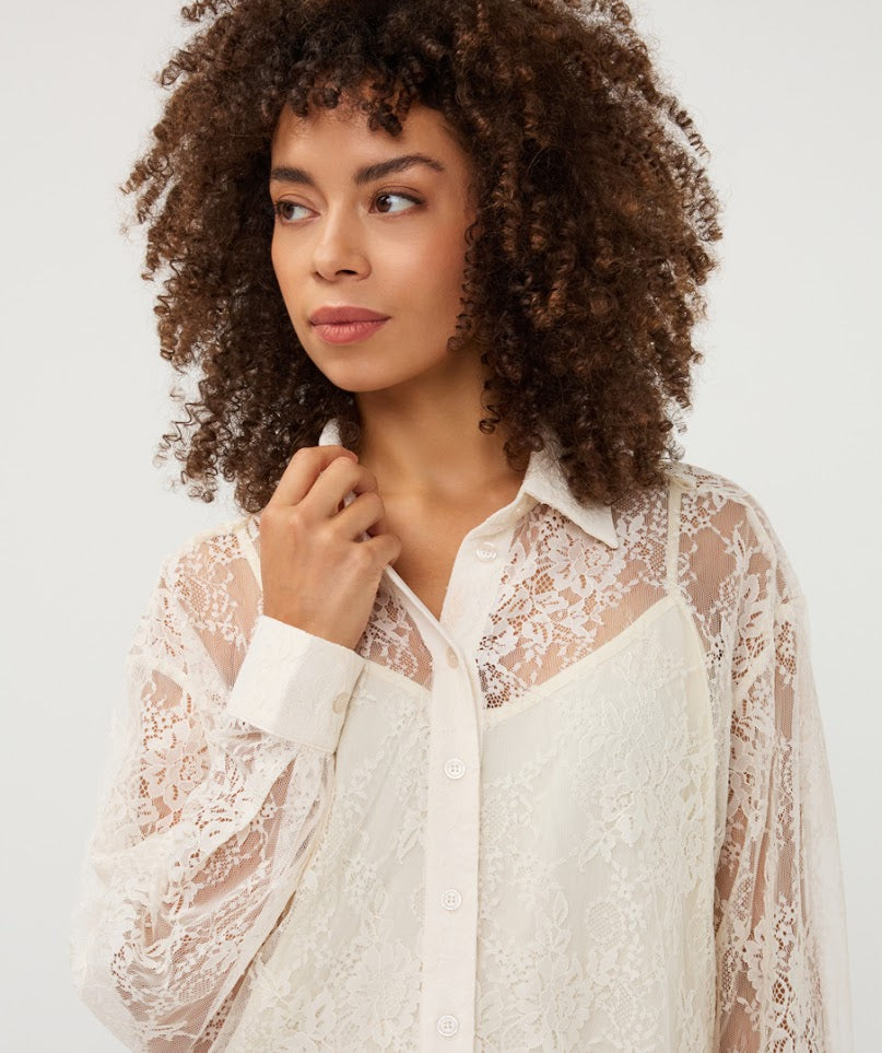 A woman with voluminous curly brown hair wearing a white lace button-up shirt over a matching white camisole, photographed against a clean light gray studio background.
