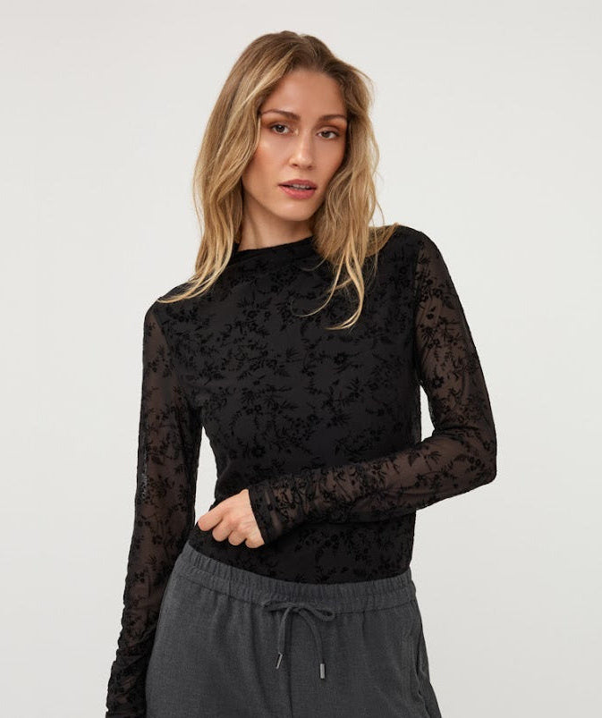 A woman modeling a black long-sleeve top with sheer floral lace sleeves and a textured black lace bodice, paired with dark gray drawstring pants, against a plain light gray studio background.